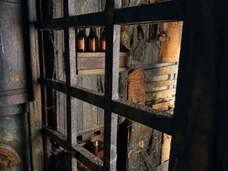 Blick durch ein Holzgitter auf Flaschen, ein verrostetes Kästchen, ein Fass und ein Fernrohr